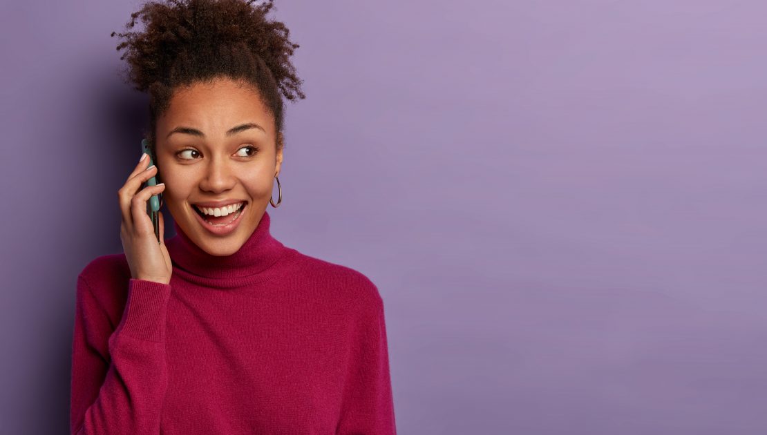 Pleased black woman has telephone conversation, looks aside, discusses pleasant news with close friend, calls someone, wears burgundy turtleneck, isolated over purple wall, copy space. Technology