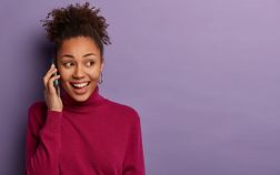 Pleased black woman has telephone conversation, looks aside, discusses pleasant news with close friend, calls someone, wears burgundy turtleneck, isolated over purple wall, copy space. Technology